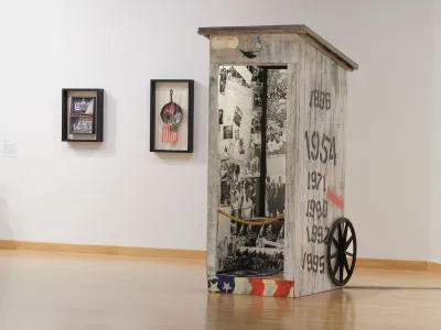 Two mixed-media artworks hang on a wall behind a sculpture that resembles an outhouse that has been decoupaged with photographs from the civil rights movement. 