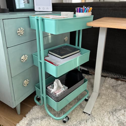 A three-tiered teal rolling cart is positioned between a light blue dresser and a wooden desk, stocked with office supplies.