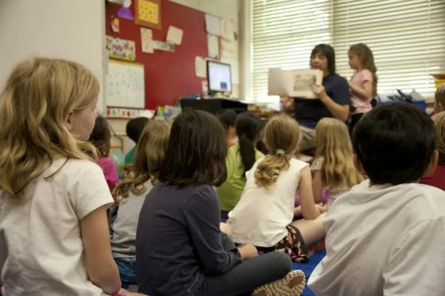 A group of young children sits cross-legged on the classroom floor, attentively listening to a teacher who is reading a picture book aloud.