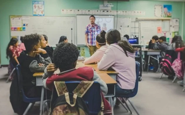 Middle-grade students sit in groups at desks, attentively facing a teacher standing at the front of the classroom near a whiteboard and projector screen